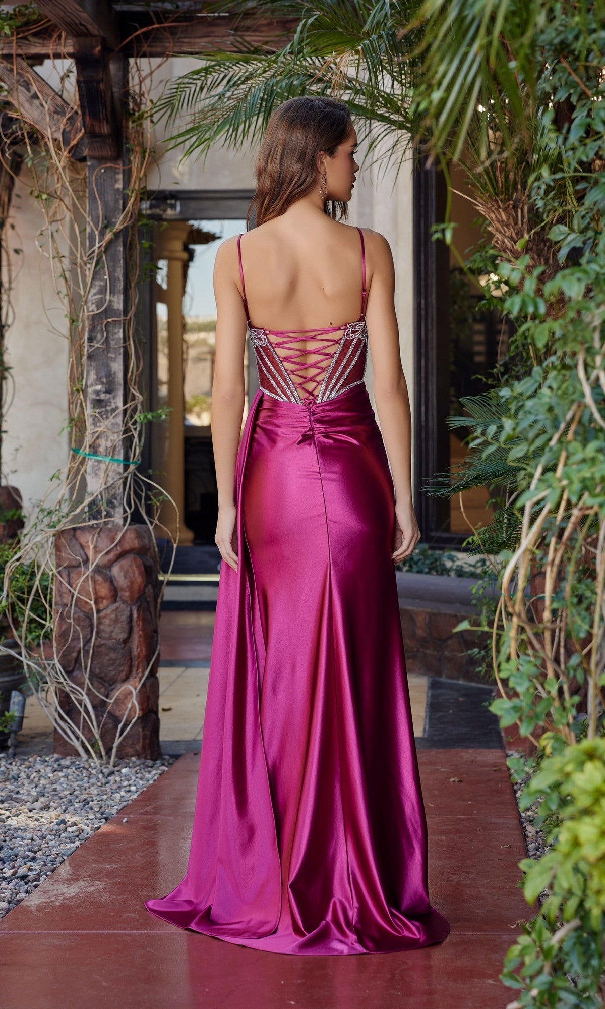 A woman stands outdoors, facing away from the camera, wearing the Nox Anabel G1788 long prom dress in magenta satin with spaghetti straps, an open lace-up back, and a floor-length skirt.